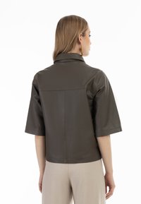 Giacca in pelle marrone con colletto, maniche corte e forma strutturata. Texture liscia e design minimalista, abbinata a pantaloni beige.