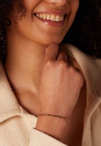 Gouden kettingarmband gedragen om een pols, met onderling verbonden ovale schakels, tegen een gestructureerde beige stoffen achtergrond.