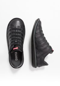 Black leather sneakers with elastic laces, textured upper, molded rubber sole, and red logo accents on the tongue and heel.