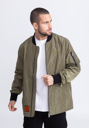 Olivgrüne Bomberjacke mit Reißverschluss vorne, gerippten schwarzen Bündchen und Kragen sowie einem Logodetail. Verfügt über eine seitliche Tasche mit Reißverschluss.