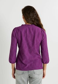 Blouse en dentelle violette avec un col à volants et des manches trois-quarts. Motif floral texturé ; associée à un pantalon gris clair légèrement ajusté.