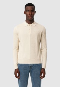 Uomo che indossa un maglione polo a maniche lunghe beige e jeans blu, in piedi contro uno sfondo grigio chiaro uniforme.