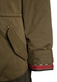 Manche de veste marron avec poche à rabat et poignet présentant une bordure à motifs rouges et noirs, gros plan sur la texture du tissu et les détails de la couture.