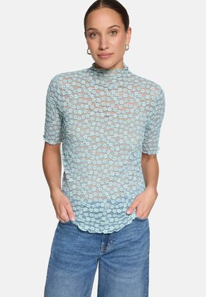 Kobieta w jasnoniebieskiej kwiecistej koronkowej bluzce z krótkim rękawem i wysokim kołnierzem, w połączeniu z niebieskimi dżinsami, ręce w kieszeniach.