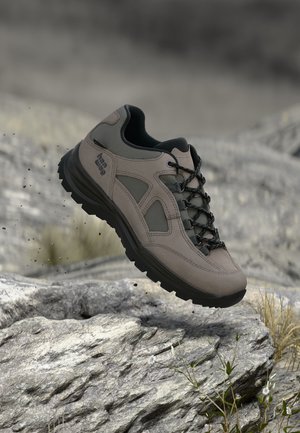 Une chaussure de randonnée grise, avec des lacets noirs et une semelle robuste, semble suspendue au-dessus d’un sentier de montagne rocailleux.