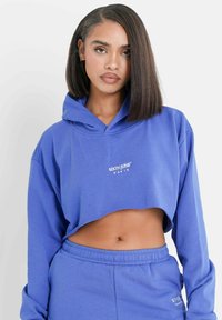 Geknipte blauwe hoodie met een capuchon en lange mouwen, voorzien van een witte bedrukte logo. Joggingbroek in bijpassende kleur maakt de outfit compleet.