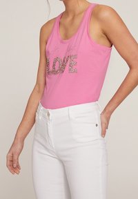 Vaaleanpunainen puuvillatoppi, jossa on kimalteleva "LOVE" grafiikka, yhdistetty valkoisiin tiukkoihin farkkuihin. Topissa on rennon istuvuuden ja leveät olkaimet.