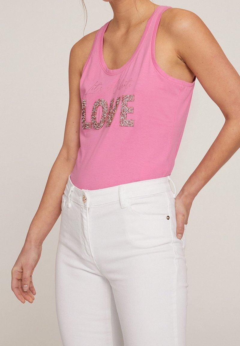 Vaaleanpunainen puuvillatoppi, jossa on kimalteleva "LOVE" grafiikka, yhdistetty valkoisiin tiukkoihin farkkuihin. Topissa on rennon istuvuuden ja leveät olkaimet.