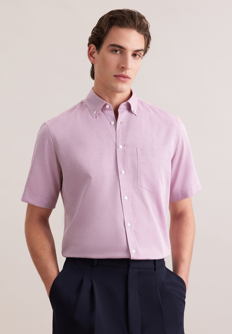 Jeune homme portant une chemise à manches courtes rose pastel avec une poche poitrine, rentrée dans un pantalon plissé bleu marine foncé.