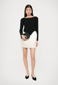 Top preto de manga longa com um decote amplo, combinado com uma saia mini creme com um design drapeado. O modelo segura uma clutch preta.