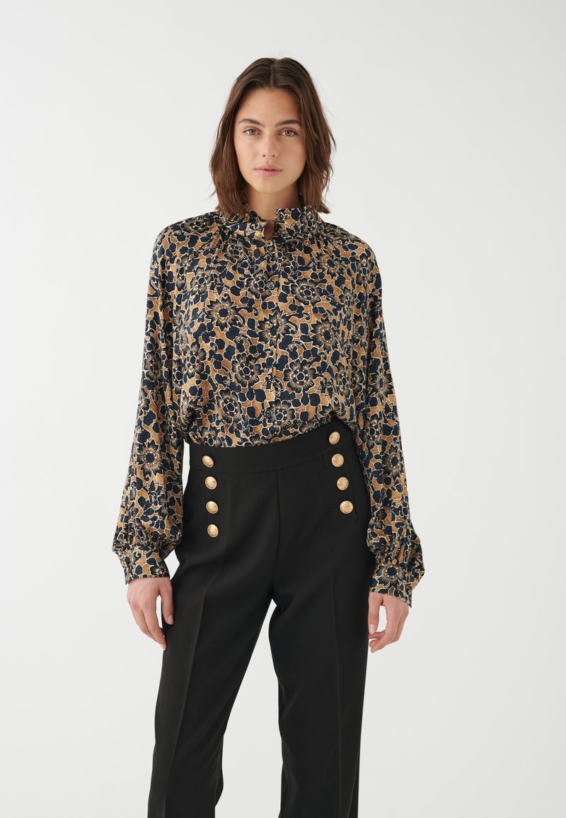 Blusa floral en tejido dorado, azul marino y negro, con un alto cuello y mangas largas y fruncidas. Pantalones negros de cintura alta con botones dorados.