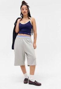 Jonge vrouw met dreadlocks, gekleed in een marineblauwe tanktop, lichtgrijze wijde shorts, witte sokken en burgundy instappers, met een zwarte jas over haar schouder.