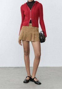 Sweat à capuche rouge côtelé zippé, mini-jupe plissée en velours côtelé marron et chaussures Mary Jane noires. Le modèle tient un petit sac à main noir.