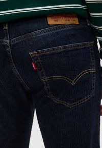 Άτομο που φοράει σκούρα μπλε τζιν Levi's 502 με ορατέ ς ραφές στην πίσω τσέπη και δερμάτινη ετικέτα μεγέθους, συνδυασμένο με πράσινο ριγέ πουκάμισο.