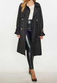Trench-coat noir à boutonnage double avec des boutons dorés et des poignets ceinturés. Porté sur un haut clair et associé à un pantalon noir en similicuir.
