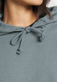 Sudadera con capucha en tejido azul verdoso apagado con una textura suave. Cuenta con un cierre de cuerda con extremos anudados para el ajuste.