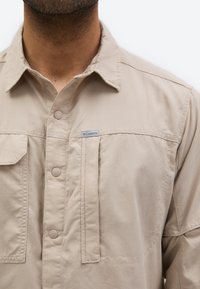 Άνδρας που φοράει μπεζ πουκάμισο Columbia με κουμπιά μπροστά και τσέπες στο στήθος, ορατός από τον λαιμό έως το πάνω μέρος του κορμού πάνω σε λευκό φόντο.