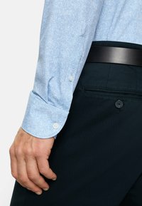 Hellblaues Fischgrätenmuster-Hemd mit Manschette und Knopf, kombiniert mit dunklen Hosen, die über eine geknöpfte Gesäßtasche verfügen. Glatte Stofftextur.