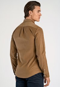 Maglietta marrone a maniche lunghe con colletto, caratterizzata da una vestibilità slim e polsini con bottoni. Tessuto liscio con una leggera lucentezza, dettagli con pince sul retro.