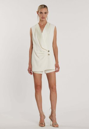 Donna bionda che indossa un abito blazer bianco senza maniche con bottoni dorati e tacchi alti dorati con cinturini, in piedi davanti a uno sfondo semplice.