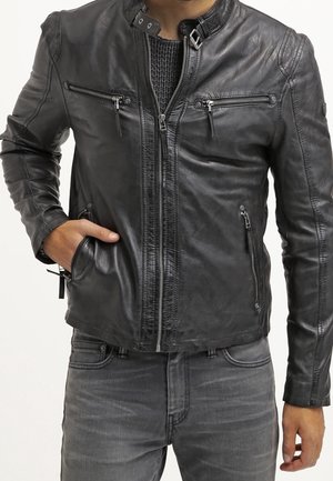 Mężczyzna w czarnej skórzanej kurtce z wieloma zapinanymi na zamek kieszeniami, w ciemnoszarych dżinsach oraz czarnym dzianym swetrze pod spodem.