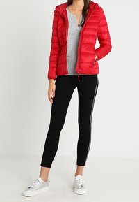 Femme portant une doudoune rouge, un pull gris, des leggings noirs avec des bandes blanches sur le côté et des baskets argentées, se tenant devant un fond uni.