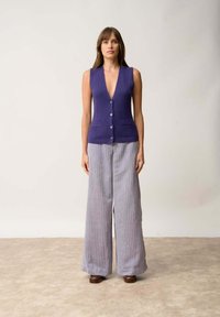 Fialová bezrukávová vesta s hlubokým výstřihem do V a dvěma předními kapsami, kombinovaná s vysokými širokými pruhovanými kalhotami v šedé a bílé barvě.