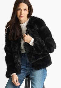 Black faux fur jacket with a textured finish, cropped design, and wide sleeves, paired with a light sweater and blue jeans.
