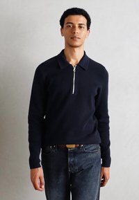 Marineblå strikkegenser med glidelås i kragen og lange ermer, kombinert med mørk denim jeans. Stoffet ser mykt og strukturert ut.