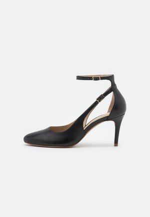 Chaussure à talon haut en cuir noir avec un bout pointu, des brides de cheville à double boucle et un détail découpé sur les côtés, sur fond blanc.