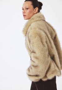 Umelý kožuch béžovej farby so širokým golierom a mäkkým, textúrovaným povrchom. Obsahuje spadnuté ramená a voľný strih.