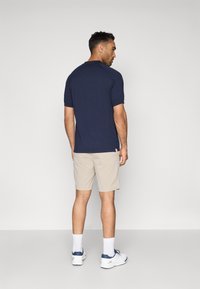 Μπλε κοντομάνικο πόλο πουκάμισο και μπεζ σορτς, με κλασική εφαρμογή. Το πόλο έχει λεία υφή και συνδυάζεται με λευκές κάλτσες και αθλητικά παπούτσια.