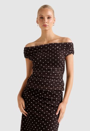 Woman wearing black off-shoulder dress with white polka dots, standing with hands resting on hips against a plain light gray background.