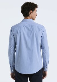 Blauw-witte gestreepte lange mouwen shirt met een button-down kraag. Heeft een rond aflopende zoom en knoopmanchetten. Achteraan is het schouderstuk zichtbaar.
