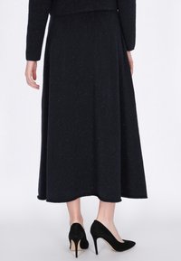 Frau in einem langen schwarzen Rock und schwarzen High Heels, die vor einem schlichten weißen Hintergrund steht.