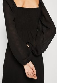 Svart klänning med fyrkantig halsringning, smockad byst och puffiga långärmar. Tyget har en texturerad yta som förstärker dess visuella detaljer.