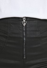 Falda de cuero negra con un cierre plateado central que presenta un tirador triangular con strass incrustados, combinada con una blusa blanca.