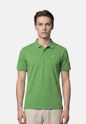 Jeune homme de face portant un polo vert à manches courtes avec un petit logo rectangulaire sur la poitrine gauche.