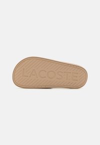 Barna gumitalp, irányított mintázattal, domború "LACOSTE" logóval és lekerekített élekkel, mely a tapadásra és tartósságra lett tervezve.