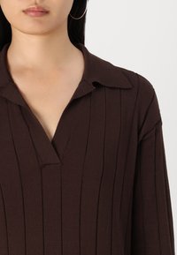 Brun ribbet strikket top med kravehals og en V-formet åbning. Stoffet har en blød tekstur og har lodrette striber.