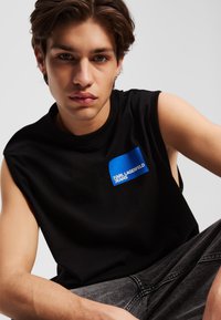 Črn brezrokavnik z okruglim izrezom, s plavo logotipno nalepko "KARL LAGERFELD JEANS". Mehka tekstura, sproščen dizajn.