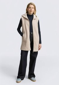 Beežist fliisvest kapuutsi ja tõmblukuga, kahel küljel taskud. Seljas musta kõrgekaelusega särgi peal ning sobitatud tumedate teksade ja spordijalanõudega.