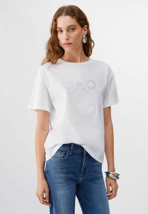 Kobieta z falującymi brązowymi włosami ubrana w biały T-shirt z ozdobnym logo, niebieskie dżinsy, duże srebrne kolczyki i srebrne bransoletki na prawej ręce.