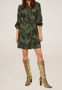 Femme portant une robe vert imprimé serpent à boutons et des bottes à talons beige imprimé serpent jusqu'au genou, se tenant devant un fond uni.