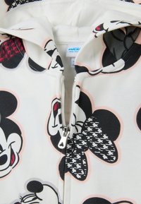 Vêtement zippé blanc avec des visages de Mickey Mouse en noir et blanc et un motif à carreaux houndstooth près de la fermeture éclair.