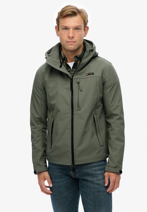 Uomo che indossa una giacca con cappuccio verde oliva sopra una camicia a quadri e una maglietta bianca, abbinata a jeans blu, in piedi contro uno sfondo bianco.