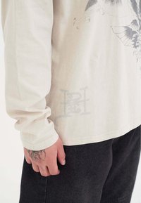 Langärmliges, hellbeiges Baumwollhemd mit Grafikdruck, mit abgerundetem Saum und Logodetail an der Seite, kombiniert mit dunkelgrauen Hosen.