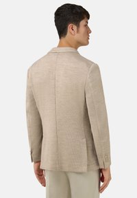 Béžový na míru šitý blazer s hladkým povrchem, jedním zadním ventilem a manžetami se čtyřmi knoflíky. Obsahuje klopové límce a boční kapsy.