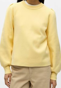 Světlounce žlutý pulovr se zaobleným výstřihem, dlouhými nafouklými rukávy a texturovanou pleteninou, v kombinaci s béžovými kalhotami.