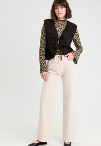 Gilet lavorato a maglia nero con un nodo frontale, sopra una maglietta a maniche lunghe con stampa leopardata verde, abbinato a jeans larghi di colore bianco sporco e scarpe nere.